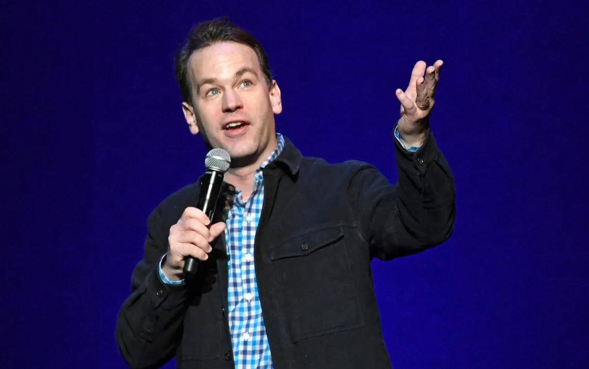 A stand-up comedian in a black jacket holds a microphone, preparing to entertain the audience with his performance.