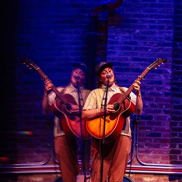 A person playing an acoustic guitar on a stage with a brick wall background. The person appears to be singing into a microphone. There is a visual effect that creates a double image of the person, making it look like there are two overlapping figures. The lighting is predominantly blue, adding a dramatic effect to the scene.