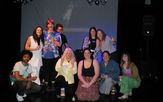 "A group of people posing together in a dimly lit room or venue. There are nine individuals in the group, with some standing and others kneeling or crouching in front. The background features a projection screen with an indistinct image on it, and there are speakers visible on either side of the group. The people are dressed in casual clothing, and some are holding drinks.