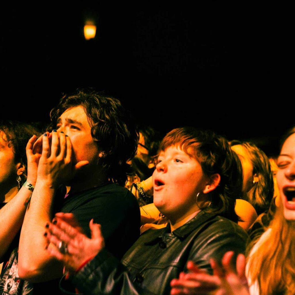 A group of people cheering for a concert at the Englert Theatre. Some individuals are clapping while others appear to be cheering or shouting. The dim lighting and focused stage lights suggest an indoor venue, creating a lively and energetic atmosphere.