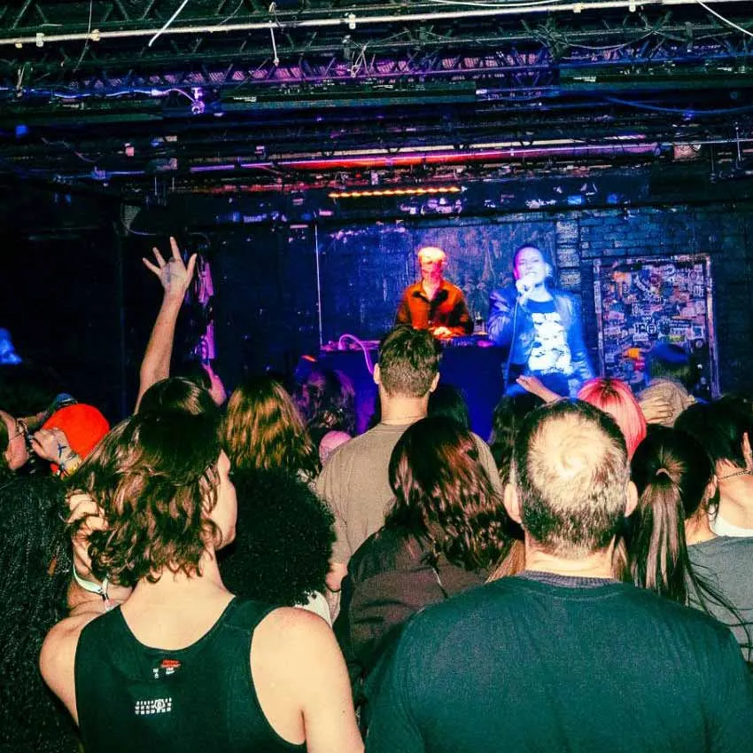 A crowded concert or live music event. The audience is facing a stage where two performers are present—one standing behind a DJ setup and the other holding a microphone. The venue appears to be a small, possibly underground club with a low ceiling and exposed pipes. The audience is engaged, with some people raising their hands. The lighting is dim with colorful stage lights illuminating the performers